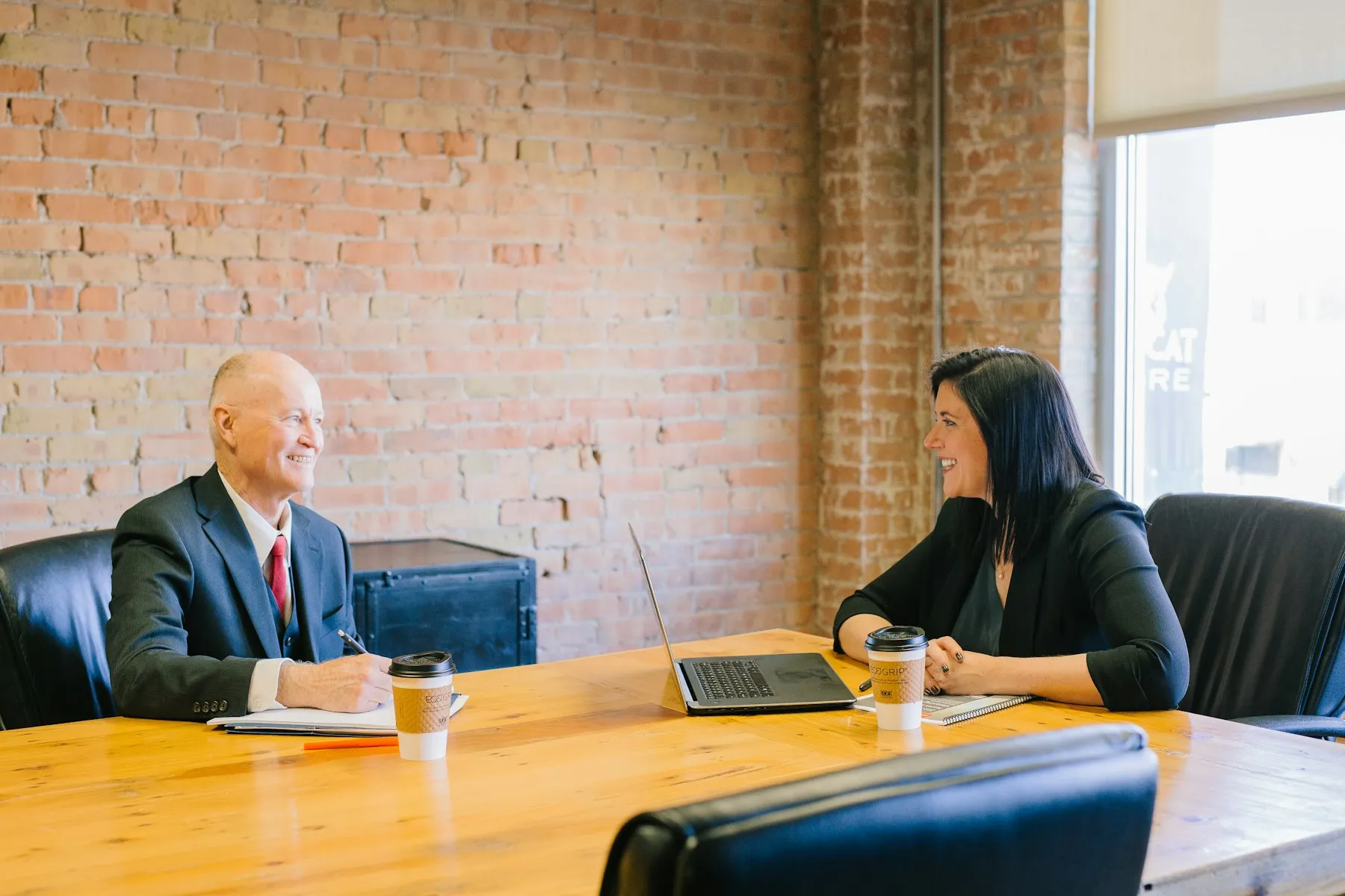 Two people talking and smiling across a table with laptops and coffee
